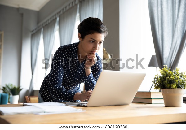 Thoughtful serious young indian ethnic woman\
student freelancer working studying on laptop computer looking at\
pc screen focused on thinking solving online problem doing research\
at home office desk.