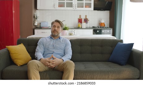 Thoughtful Serious Young Man Alone Sitting Alone On Sofa At Home. Bored And Pensive Male Alone At Home Looking At Camera. Uncertainty Concept.