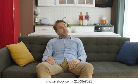 Thoughtful Serious Young Man Alone Sitting Alone On Sofa At Home. Bored And Pensive Male Alone At Home Looking At Camera. Uncertainty Concept.