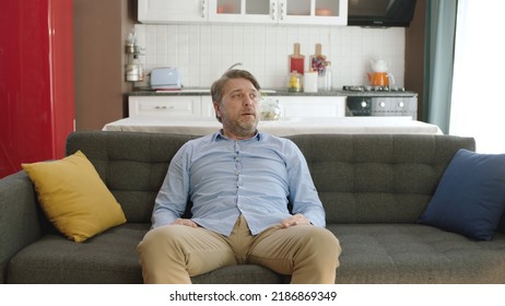 Thoughtful Serious Young Man Alone Sitting Alone On Sofa At Home. Bored And Pensive Male Alone At Home Looking At Camera. Uncertainty Concept.