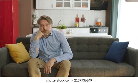 Thoughtful Serious Young Man Alone Sitting Alone On Sofa At Home. Bored And Pensive Male Alone At Home Looking At Camera. Uncertainty Concept.