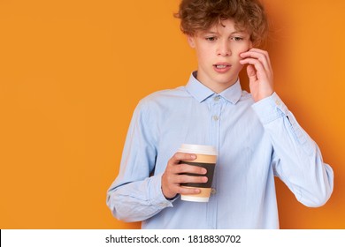 thoughtful sleepy boy has no enough sleep, need sleep in the morning before going at school, hold cup of coffee in hands, looks down. orange background
