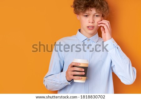 thoughtful sleepy boy has no enough sleep, need sleep in the morning before going at school, hold cup of coffee in hands, looks down. orange background