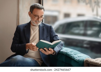 Thoughtful smart caucasian senior man with glasses engrossed in reading a book, comfortably seated near a window in a cozy corner with soft natural light, enjoy spare time