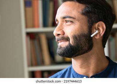 Thoughtful smiling bearded indian ethnic business man relaxing wearing earbud thinking listening to music, business training podcast or audio book in mobile application. Close up portrait. Copy space