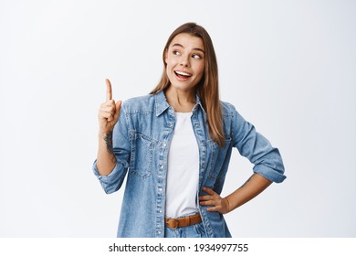 Thoughtful smiling woman with fair hair, raising finger and pointing up, having good point or idea, showing advertisement, standing against white background