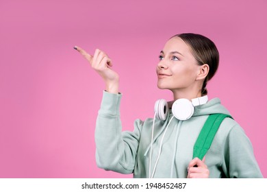 Thoughtful Smiling Woman, A Student About To Study, With A Backpack And With Headphones. Portrait On A Solid Monochrome Pink Background. The Female Shows With Her Index Finger, Copy Space