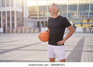 Thoughtful sportive mature man in sportswear standing outdoors with basketball ball, ready for workout