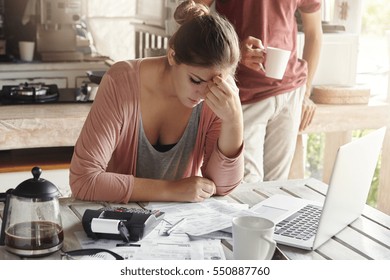 Thoughtful Stressed Young Female Sitting At Kitchen Table With Papers And Laptop Computer Trying To Work Through Pile Of Bills, Frustrated By Amount Of Domestic Expenses While Doing Family Budget