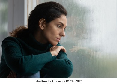 Thoughtful stressed young hispanic latin woman sitting on windowsill, looking outside on rainy weather, having depressive or melancholic mood, suffering from negative thoughts alone at home.