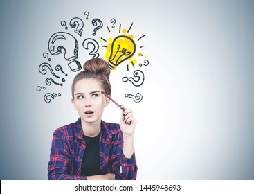 Thoughtful teen girl in casual clothes sitting near gray wall with question marks and lightbulb drawn on it. Concept of good idea. Mock up