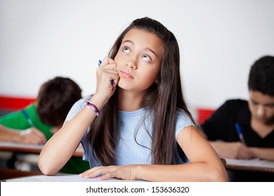 Thoughtful teenage schoolgirl looking up while sitting at desk during examination with friends in background - Powered by Shutterstock