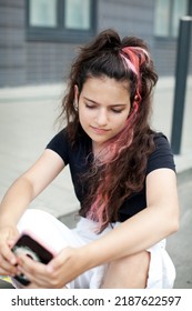 Thoughtful Teenager Girl With Long Dark Curly Colored Hair Dyed Pink Looks Into Smartphone Gadget, Chats Or Video Calls