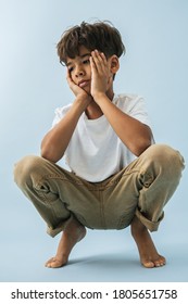 Thoughtful Tired Barefoot Indian Asian Kid Sitting Low, Knees Up, Hands On Cheeks. Over Blue Background.