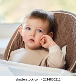 A Thoughtful Toddler Baby Sits At An Empty Table Without Food. A Child With A Doubting Face On A High Children Chair. Kid Aged One Year And Two Months