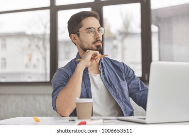 Thoughtful unshaven male journalist tries to write creative article, works on laptop computer, drinks coffee from paper cup. Attractive young man at workplace, thinks about creating new project - Shutterstock ID 1006653511
