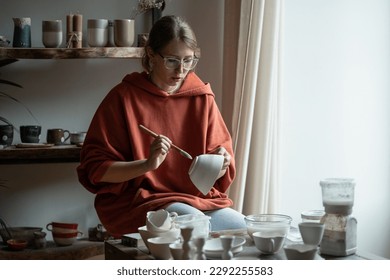 Thoughtful woman artist varnishing pottery made from natural clay. Creation porcelain products for home. Focused girl sits at desk in workshop in process art therapy to restore psychological health - Shutterstock ID 2292255583