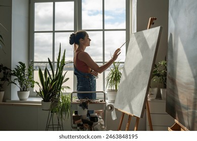 Thoughtful woman artist working on painting. Girl outstretched hand with brush reaches for canvas, making careful thoughtful strokes, creating masterpieces. Working process in art gallery, hobby.  - Powered by Shutterstock