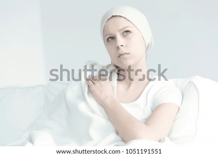 A thoughtful woman with cancer sitting under a blanket in a white room
