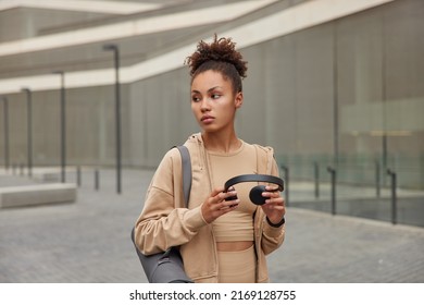 Thoughtful woman with curly combed hair looks away pensively holds stereo headphones dressed in beige sportswear carries rolled karemat poses outdoors against blurred background. Active lifestyle