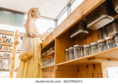 Mujer reflexiva con el tarro de cristal vacío y los limones frescos en bolso de la malla que compra productos sin el empaquetado plástico en la tienda cero de la basura. Cliente femenina hace compras conscientes en tienda gratuita de plástico moderno.