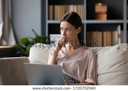Thoughtful woman holding laptop on laps, pondering ideas or tasks, sitting on couch at home, dreamy young female touching chin and looking in distance, lost in thoughts, waiting for message