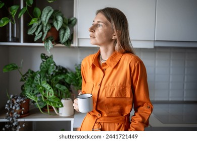 Thoughtful woman holding mug standing in kitchen surrounded by houseplants. Portrait of calm pensive confident female in orange dress holds tea cup, thinking, looking at window.  - Powered by Shutterstock