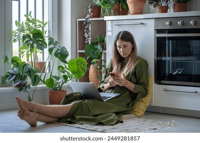 Thoughtful woman with laptop on knees looking at screen cellphone sits comfortably on floor among indoor plants at home. Focused freelancer female remote works, uses smartphone and portable computer. - Powered by Shutterstock