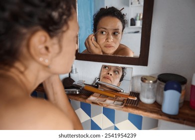 A thoughtful woman looks at her reflection while applying makeup, surrounded by toiletries in the bathroom. The image conveys contemplation, beauty routine, and self-care. - Powered by Shutterstock