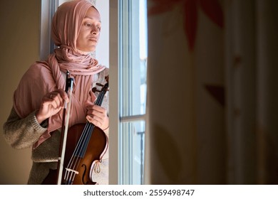 A thoughtful woman in a pink hijab stands by a window, holding a violin and bow, bathed in sunlight. Her expression suggests contemplation and introspection, creating a serene atmosphere. - Powered by Shutterstock