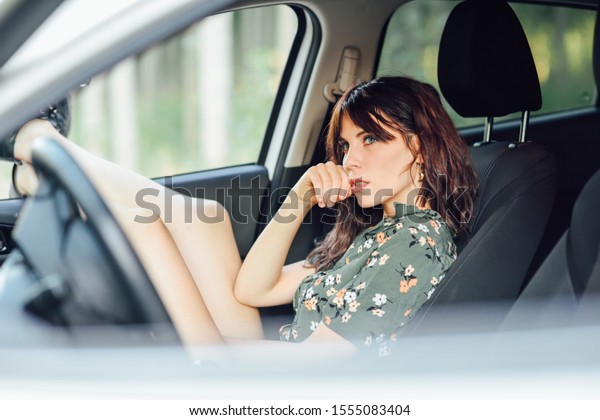 Thoughtful\
woman resting in a white car pulling her feet out the window.\
Female wearing flowered shirt and\
shorts.