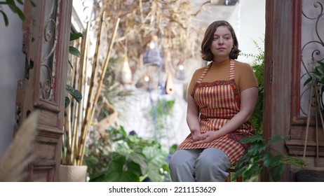 Thoughtful Woman Woman Sitting At Stool Inside Floriculture Store. Pensive Female Employee Resting Inside Local Business Shop In Contemplation