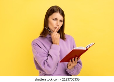 Thoughtful woman standing with paper notebook and pen, looking away with pensive expression, making to do list, wearing purple hoodie. Indoor studio shot isolated on yellow background.