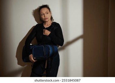 A thoughtful woman stands in dramatic lighting, clutching a gym bag. The shadows create a contemplative, moody atmosphere, suggesting themes of introspection and preparation. - Powered by Shutterstock