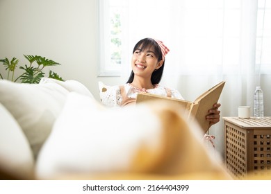 Thoughtful woman student wearing turban and holding a book. Girl studying at home on beige couch, wrapped up in white blanket on sofa with positive thinking