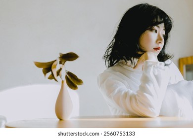 Thoughtful Woman in White Blouse Sitting by a Table with a Minimalist Vase and Greenery, Capturing Serenity and Contemplation in Natural Light. - Powered by Shutterstock