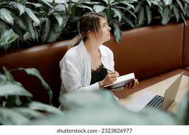 Thoughtful woman in white shirt and black lace dress lost in thought, looking away while writing notes in diary. Freelancer sitting on couch at table in front of laptop surrounded by plants. - Powered by Shutterstock