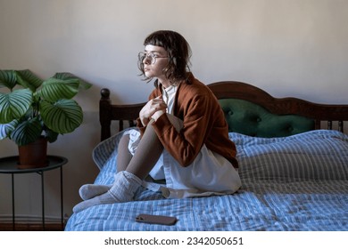 Thoughtful worried teen girl sits on bed sadly look at window waiting call, important message on smartphone. Lonely upset young woman holding knees in hand thinking about problem received bad news  - Powered by Shutterstock