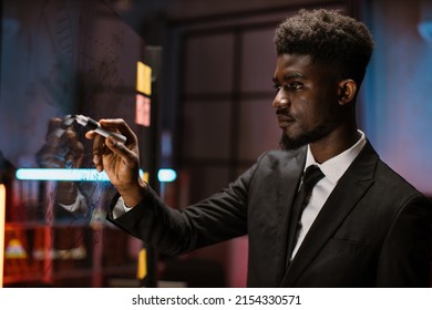 Thoughtful Young African American Business Man Professional Expert, Standing In Front Of Glass Wall With Paper Stickers And Looking On Project Plans, Working At His Office Late In The Evening.