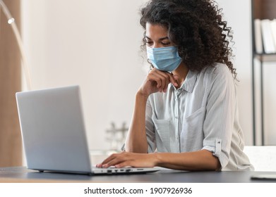A thoughtful young African American businesswoman with curly hair wearing a protective face mask sitting at the desk and working on a laptop, back to the office after covid-19 lockdown concept