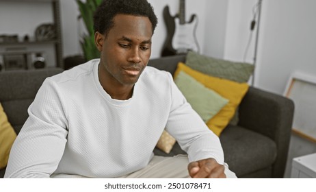 A thoughtful young african american man wearing a white sweater, sitting indoors in a well-decorated living room. - Powered by Shutterstock