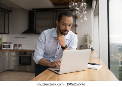 Thoughtful young african male stand by desk with laptop at home kitchen read important message email think on answer create respond. Focused biracial man student contact friend using messenger pc app