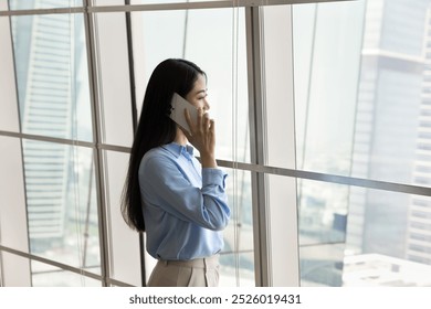 Thoughtful young Asian business woman talking on cellphone in office space, standing at large window, looking away at city view, thinking on conversation, commercial communication - Powered by Shutterstock