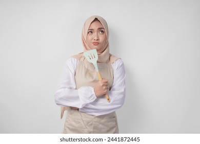 Thoughtful young Asian Muslim woman in hijab and cream apron holding spatula and kitchen cooking utensils, thinking hard what food menu to cook - Powered by Shutterstock