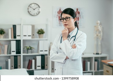 thoughtful young asian woman doctor standing in clinic office. girl medical staff with hand on chin thinking solution to solve problem. pensive female nurse serious considering cross arms in hospital