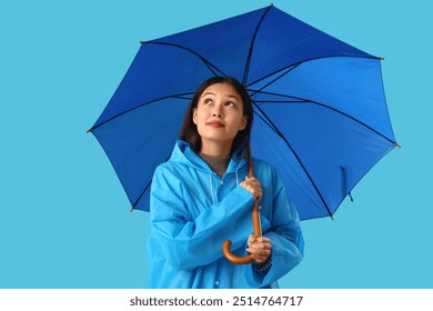 Thoughtful young Asian woman in raincoat with umbrella on blue background