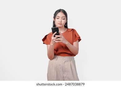 Thoughtful young Asian woman wearing brown shirt looking at her smartphone with serious expression, isolated by white background. - Powered by Shutterstock