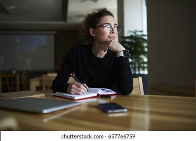Thoughtful Young Author Businesswoman And Thinking On Writing Interesting Article.Woman In Stylish Eyeglasses With Good Lenses Look Out Of Window And Pondering New Plans While Noting Them In Notepad