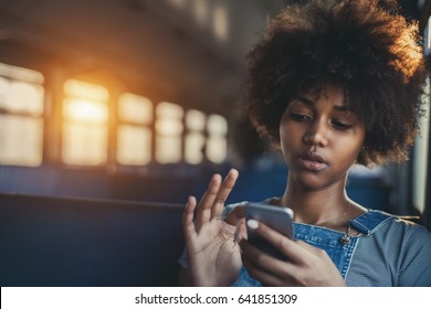 Thoughtful young biracial girl using smart phone while sitting alone in suburban train, curly african teenage female having online chat with her sister in train with copy space for your text or logo - Powered by Shutterstock