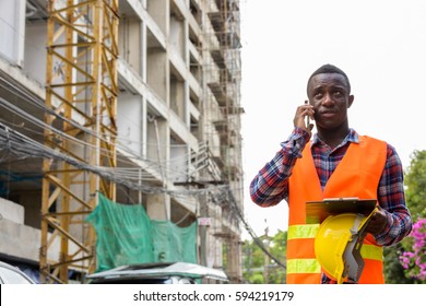 Thoughtful Young Black African Man Construction Worker Talking On Mobile Phone While Holding Safety Helmet And Clipboard At Building Site